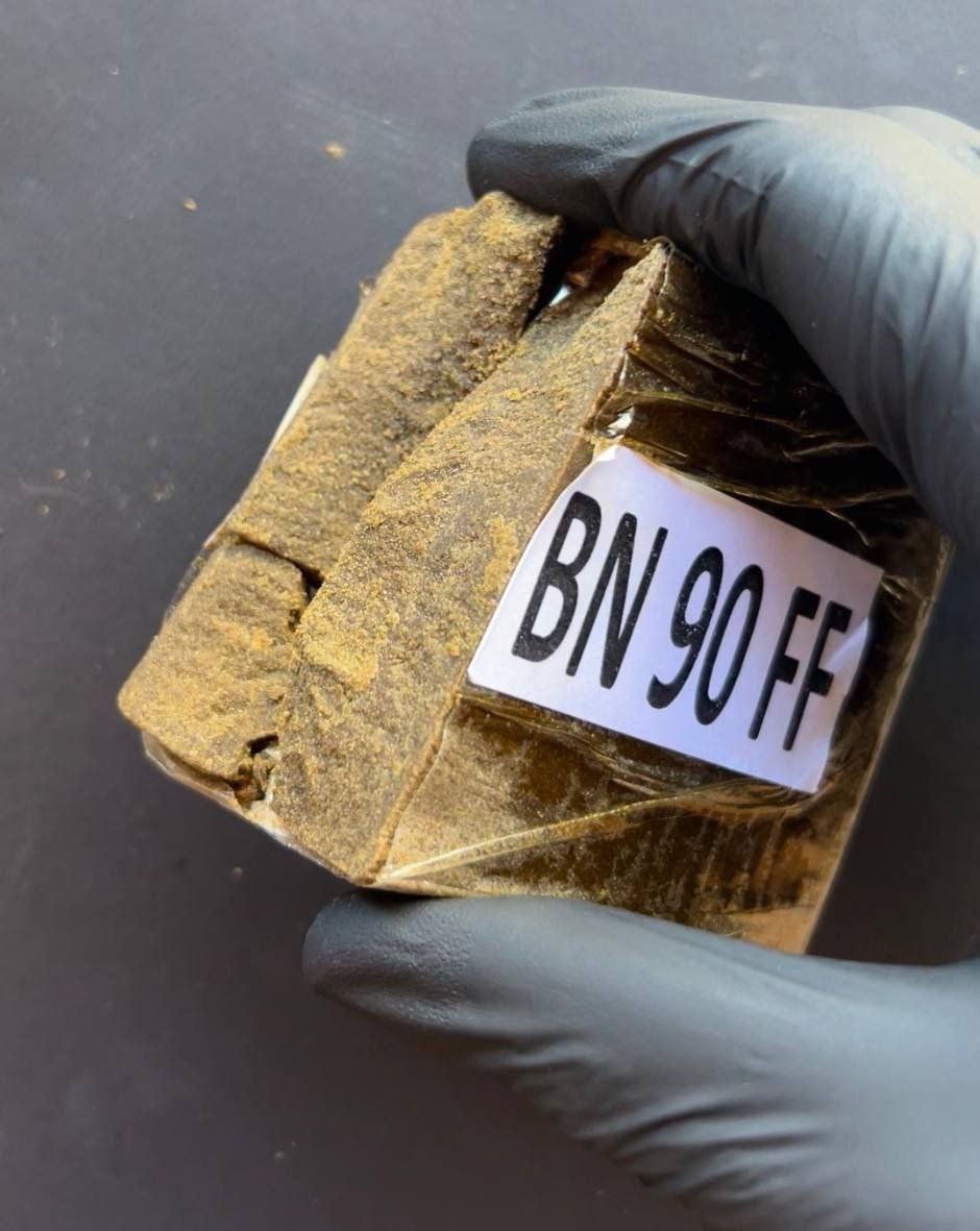 Gloved hand holding a stamped BN 90 FF hash brick wrapped in film against a dark background