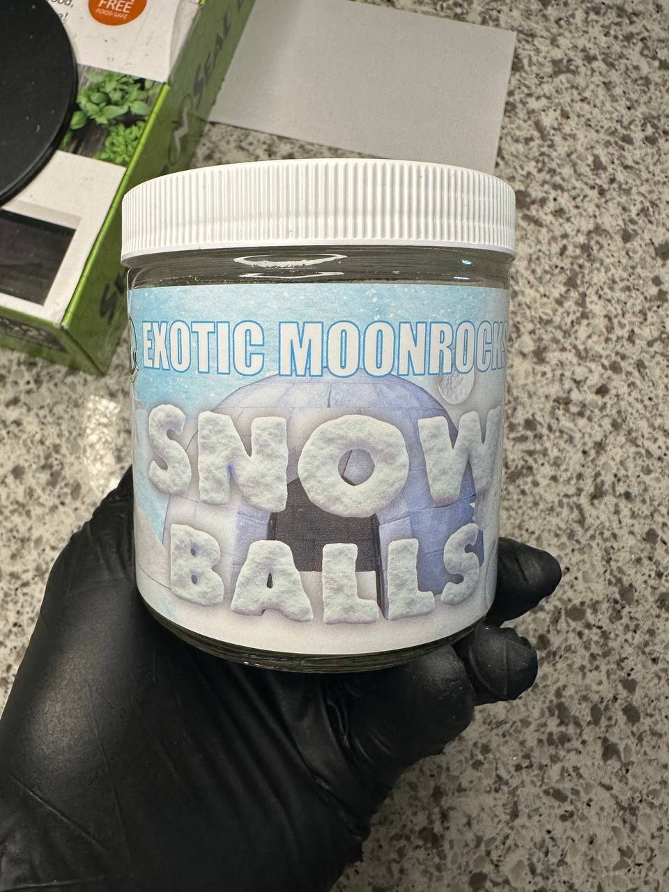 Black-gloved hand holding a jar of Exotic Moonrocks Snow Balls with the branded label facing the camera